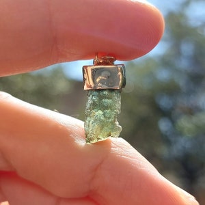 May include: A green gemstone pendant with a gold setting. The gemstone is a rough, natural shape and has a green and gold color.