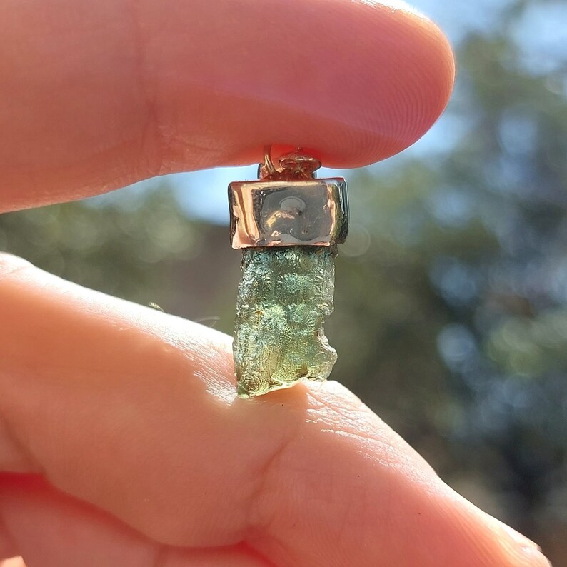 May include: A green gemstone pendant with a gold setting. The gemstone is a rough, natural shape and has a green and gold color.