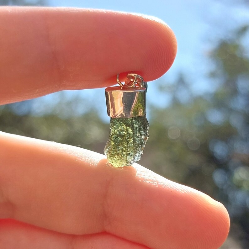 May include: A green and silver pendant featuring a raw, uncut green gemstone. The gemstone is set in a silver bezel and has a silver bail for attaching to a chain.