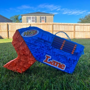 May include: A blue and orange Nerf gun shaped piñata with the name "Lane" written on it. The piñata is sitting on a green grassy lawn.
