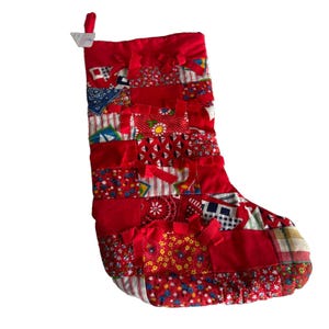 May include: A Christmas stocking constructed from a patchwork of various red-toned fabrics, including floral, gingham, and solid patterns. The stocking features horizontal fabric strips with frayed edges and a small fabric loop at the top corner.