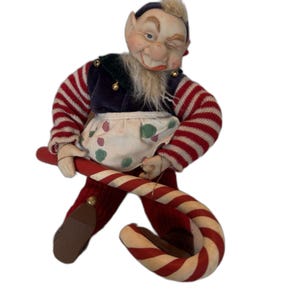 Könnte beinhalten: Eine bewegliche Elfenpuppe mit zwinkerndem Gesichtsausdruck, bekleidet mit einer dunkelblauen Samtweste, rot-weiß gestreiften Ärmeln und einer weißen Schürze mit grünen und roten Punkten. Die Elfe hält eine große rot-weiß gestreifte Zuckerstange.