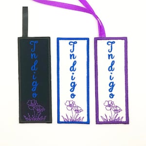 May include: Three fabric bookmarks with the word "Indigo" embroidered in different colors. The bookmarks are black, white, and purple, and each has a small purple flower design at the bottom.