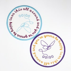 Op de afbeelding: Twee geborduurde patches met een witte achtergrond en kleurrijke stiksels. De bovenste patch is lichtblauw met de tekst "turn this all around and you're gonna be happy" en een blauw vlinderontwerp. De onderste patch is paars met de tekst "you're gonna be happy turn this all around" en een paarse vlinderontwerp. Beide patches hebben de tijd "00:00" in het midden.
