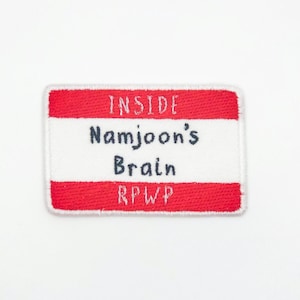 May include: A rectangular patch with a red border and the words "INSIDE Namjoon's Brain RPWP" in navy blue and white. The patch is likely made of fabric and is designed to be sewn or ironed onto clothing or accessories.