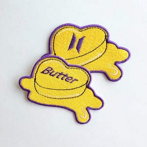 Puede incluir: Dos parches amarillos en forma de corazón con contornos morados. Un parche dice "Butter" y el otro tiene las letras "II" en morado. Los parches están diseñados para parecerse a la mantequilla derretida.