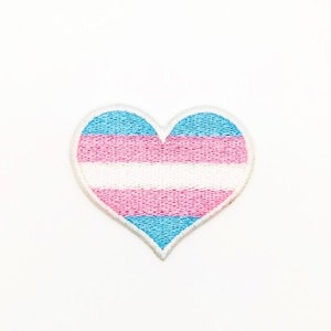 May include: An embroidered heart-shaped patch displaying the transgender pride flag. The design features horizontal stripes of light blue, pink, and white, with a white border. Ideal for attaching to fabric items. The background is white.