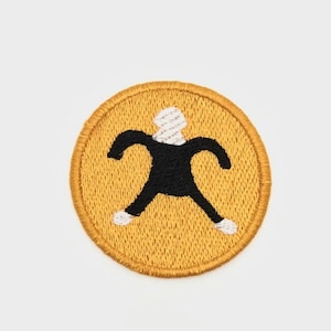 May include: A yellow embroidered patch featuring a black stick figure wearing a white hat. The stick figure has outstretched arms and legs.