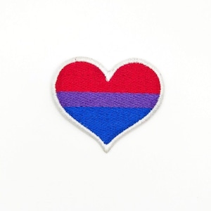 Puede incluir: Un parche blanco en forma de corazón con un diseño de bandera del orgullo bisexual rojo, morado y azul.