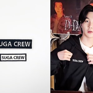 Puede incluir: Dos parches bordados en blanco y negro con el texto "SUGA CREW" sobre un fondo blanco. Una persona que lleva una sudadera con capucha negra con el texto "SUGA CREW" bordado en la parte delantera.