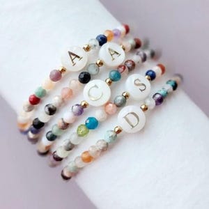 May include: Five beaded bracelets with colorful gemstones and gold accents. Each bracelet features a white circular charm with a gold letter: A, C, S, and D. The bracelets are stacked on a white surface.