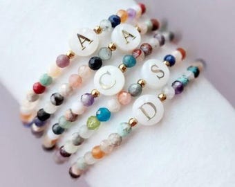 May include: Five beaded bracelets with colorful gemstones and gold accents. Each bracelet features a white circular charm with a gold letter: A, C, S, and D. The bracelets are stacked on a white surface.