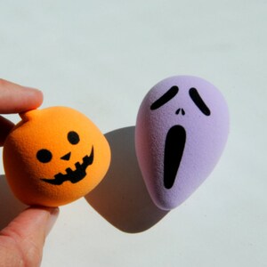 May include: Two Halloween-themed foam balls. One is orange and shaped like a pumpkin with a black jack-o'-lantern face. The other is purple and shaped like a ghost with a black screaming face.