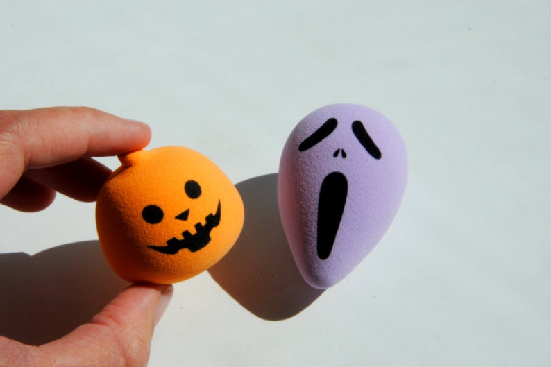 May include: Two Halloween-themed foam balls. One is orange and shaped like a pumpkin with a black jack-o'-lantern face. The other is purple and shaped like a ghost with a black screaming face.