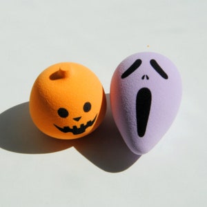 May include: Two Halloween-themed makeup sponges, one orange with a jack-o'-lantern face and one purple with a screaming ghost face.
