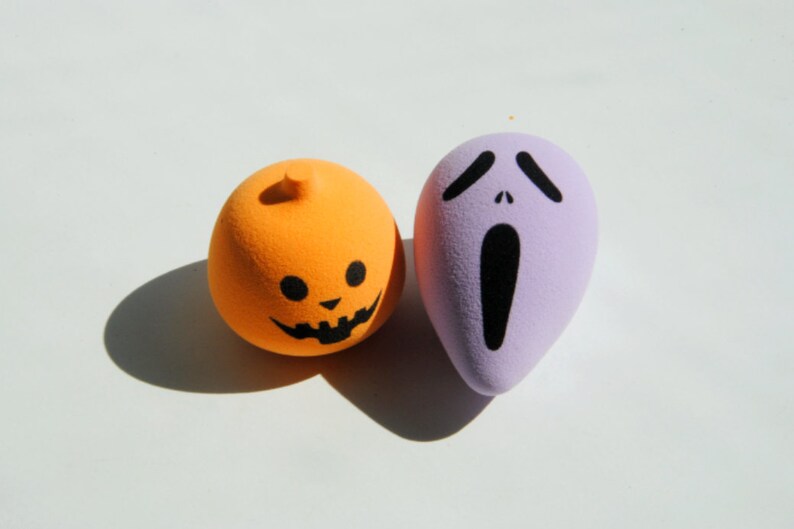 May include: Two Halloween-themed makeup sponges, one orange with a jack-o'-lantern face and one purple with a screaming ghost face.