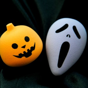 May include: A white ghost face mask with a black scream and two black eyes, next to a bright orange pumpkin with a black jack-o-lantern face.