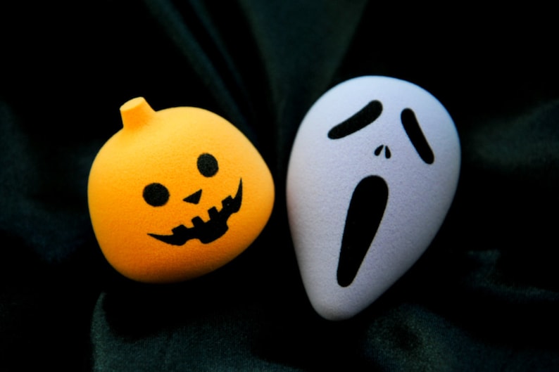 May include: A white ghost face mask with a black scream and two black eyes, next to a bright orange pumpkin with a black jack-o-lantern face.