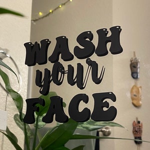 Könnte beinhalten: Ein Badezimmerspiegel mit einem schwarzen Vinyl-Aufkleber, auf dem in einer Retro-Schrift "WASH your FACE" steht. Der Spiegel spiegelt eine Pflanze und Lichterketten wider.