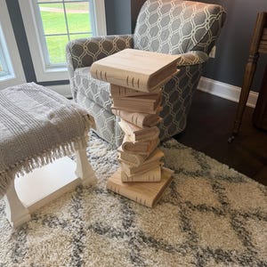 May include: A wooden side table crafted to resemble a stack of books. The table is made of light-colored wood and features a rectangular top. The base is a stack of book-shaped forms, creating a unique and decorative piece of furniture.