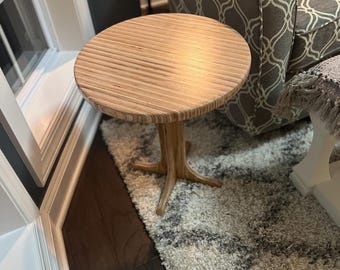 May include: A small, round wooden side table with a light brown finish. The tabletop has a ribbed texture, and the base is a tree-trunk-like design. The table is approximately 20 inches in diameter and stands on a patterned rug.