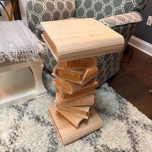 May include: A light-coloured wooden side table featuring a base and central column constructed from stacked, horizontal book-shaped wooden segments, topped with a flat, rectangular wooden surface with rounded edges.