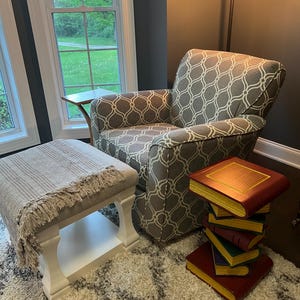 May include: A cozy reading nook featuring a patterned armchair, a matching ottoman with a fringed throw, and a stack of decorative book-shaped side tables. The room has a neutral color palette with a shaggy rug and natural light from a large window.