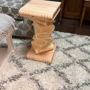 May include: A light-colored wooden side table constructed from a stack of carved book shapes. The base and top are flat, rectangular book forms, while the central support column consists of several smaller, angled book-shaped wooden pieces.