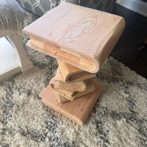 May include: A light-colored wooden side table constructed from stacked, book-shaped pieces of natural pine. The table features a square top and a base formed by several layers of wood that mimic the appearance of a vertical stack of books.