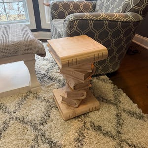 Könnte beinhalten: Ein einzigartiger Beistelltisch aus hellem Holz, der wie ein Stapel Bücher gestaltet ist. Der Tisch hat eine quadratische Oberseite und das Design beinhaltet das Aussehen von Buchseiten und -rücken, wodurch ein skurriles und künstlerisches Stück entsteht.