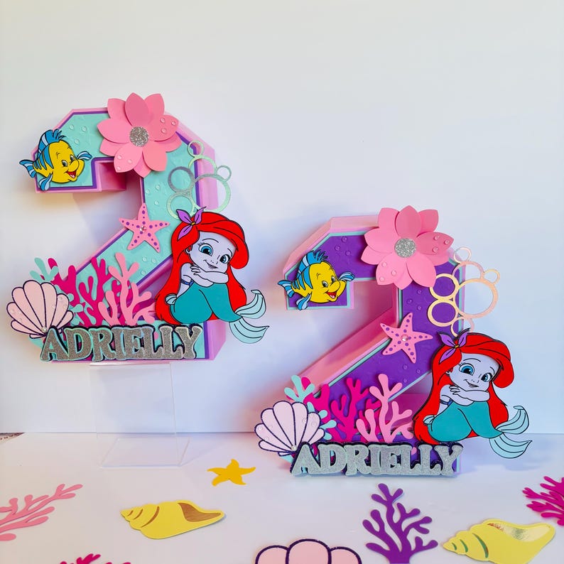 May include: Two purple and pink 3D number two cake toppers with a mermaid and Flounder character, seashells, coral, and the name Adrielly.