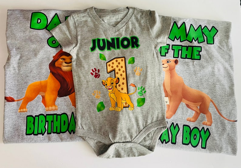 May include: A gray t-shirt and onesie set featuring Lion King characters. The t-shirts read "Daddy of the Birthday Boy" and "Mommy of the Birthday Boy". The onesie says "Junior 1" with Simba graphics and paw prints.