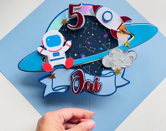 Puede incluir: Un adorno para pastel de papel azul y blanco con un cohete, un astronauta y estrellas. El número 5 está en el cohete y el nombre "Jai" está en una pancarta.