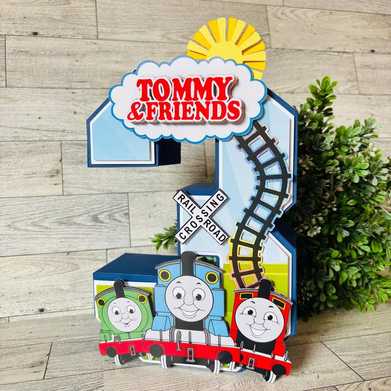 May include: A colorful, layered papercraft decoration featuring the text "TOMMY & FRIENDS" above a train scene. The design includes a blue number 2, a sun, a railroad crossing sign, and three smiling train engines in green, blue, and red.