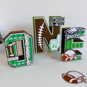 Op de afbeelding: Groene en bruine kartonnen letters die "ONE" spellen met voetbalthema-decoraties. De letter "O" heeft een voetbalveldontwerp met de woorden "1st Down" en "KAZARIS" erop. De letter "N" heeft een bruin gestikt voetbalontwerp. De letter "E" heeft een groen voetbalveldontwerp met een voetbalhelm en de woorden "TOUCH DOWN" en "EAGLES".