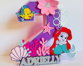 May include: A purple and pink number two cake topper with a mermaid, a fish, and a starfish. The topper also has a pink flower, coral, and seashells. The name "Adrielly" is written in silver glitter on the bottom of the topper.