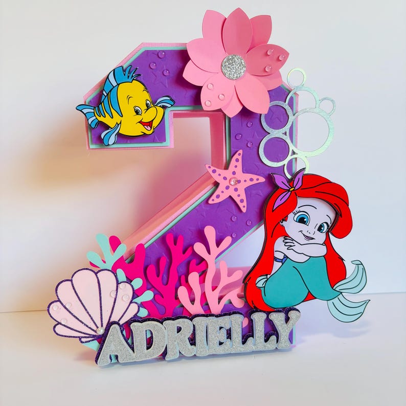 May include: A purple and pink number two cake topper featuring a mermaid, a fish, and a starfish. The topper also includes a pink flower, coral, and seashells. The name "Adrielly" is written in silver glitter at the bottom of the topper.