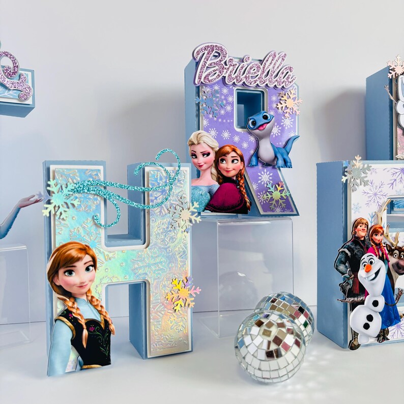 Pode incluir: Letras azul claro com detalhes brilhantes, soletrando um nome e apresentando personagens do filme "Frozen". A letra "R" tem o nome "Briella" no topo. As letras s&atilde;o decoradas com flocos de neve e imagens de Elsa, Anna, Olaf e outros personagens.