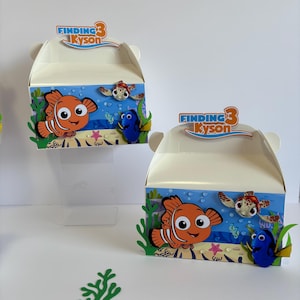 Peut inclure: Deux boîtes de faveur de fête sur le thème de Nemo avec un schéma de couleurs bleu, orange et jaune. Les boîtes présentent un personnage du film Le Monde de Nemo, Dory, et une tortue de mer. Les boîtes ont une poignée et disent "Finding 3 Kyson" sur le dessus.