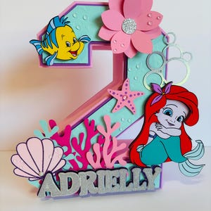 May include: A pink and blue 3D number two cake topper featuring a mermaid, a starfish, a seashell, coral, and a yellow fish. The topper also includes a pink flower and the name "Adrielly" in silver glitter.