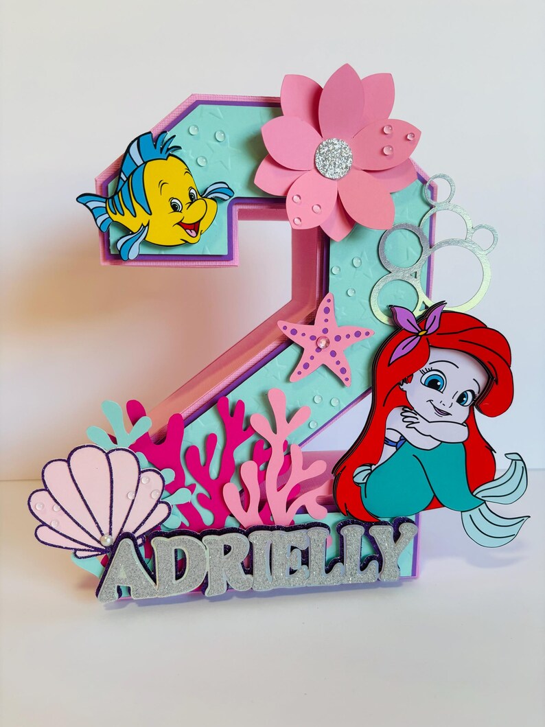 May include: A pink and blue 3D number two cake topper featuring a mermaid, a starfish, a seashell, coral, and a yellow fish. The topper also includes a pink flower and the name "Adrielly" in silver glitter.