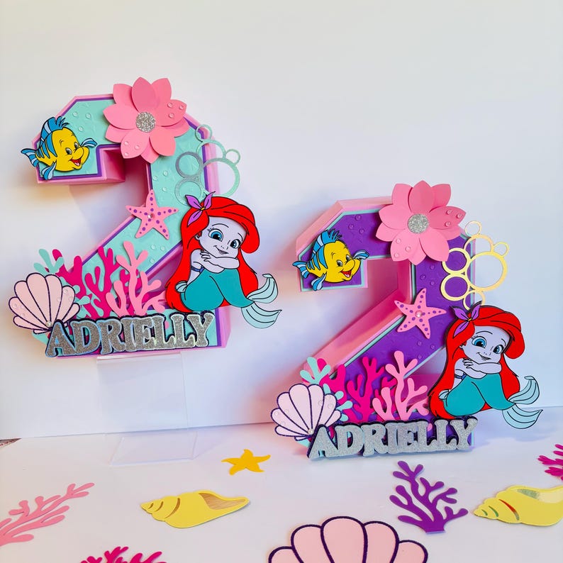 May include: Two pink and purple 3D number two cake toppers with a mermaid theme. Each number has a mermaid, seashells, coral, and a flower. The numbers are decorated with glitter and the name "Adrielly" is written on each topper.