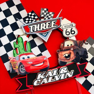 May include: A red and black cake topper featuring the characters Lightning McQueen and Mater from the Disney Pixar Cars movie. The topper also includes a checkered flag, a cactus, a Route 66 sign, and a banner that says "Kai & Calvin" with the number "Three".
