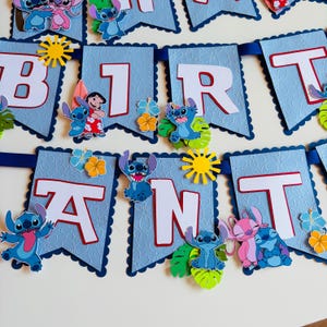Puede incluir: Una pancarta azul claro con letras blancas que deletrean "BIRTHDAY". La pancarta est&aacute; decorada con personajes de dibujos animados, flores y formas de sol. La pancarta est&aacute; ensartada en una cinta azul, ideal para una fiesta de cumplea&ntilde;os.