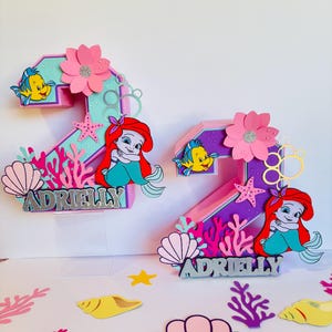May include: Two pink and purple 3D number two cake toppers featuring a mermaid, a starfish, and a seashell. The toppers are decorated with a pink flower, a yellow fish, and the name "Adrielly".