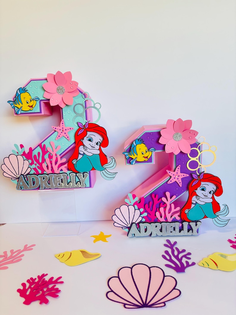 May include: Two pink and purple 3D number two cake toppers featuring a mermaid, a starfish, and a seashell. The toppers are decorated with a pink flower, a yellow fish, and the name "Adrielly".