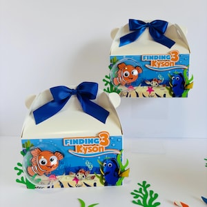 Peut inclure: Deux boîtes de faveur de fête sur le thème de Nemo à la recherche de Nemo avec des nœuds de ruban bleu. Les boîtes sont blanches avec un design bleu, orange et jaune mettant en scène Dory et Nemo. Le texte sur les boîtes indique "Finding 3 Kyson".