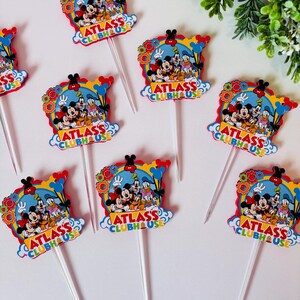 Peut inclure: Plusieurs d&eacute;corations de cupcakes color&eacute;es avec Mickey Mouse, Donald Duck et Dingo. Chaque d&eacute;coration a un fond bleu avec le texte "ATLASS CLUBHOUSE" et un b&acirc;ton en plastique transparent.