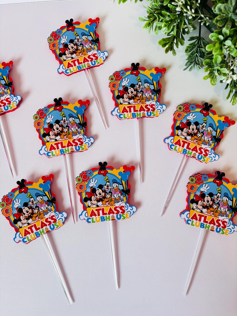 Peut inclure: Plusieurs d&eacute;corations de cupcakes color&eacute;es avec Mickey Mouse, Donald Duck et Dingo. Chaque d&eacute;coration a un fond bleu avec le texte "ATLASS CLUBHOUSE" et un b&acirc;ton en plastique transparent.