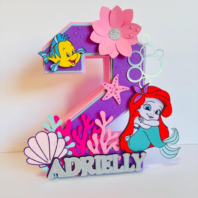 May include: A purple and pink 3D number two cake topper featuring a mermaid, a starfish, coral, seashells, and bubbles. The topper also includes a yellow fish with a blue tail and the name "ADRIELLY" in silver glitter letters.